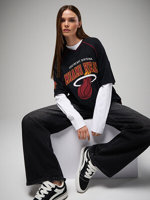 Modelka v černé košili Miami Heat s logem týmu, s bílým dlouhým rukávem pod ní, v černých džínách a sportovních botách na šedém pozadí.