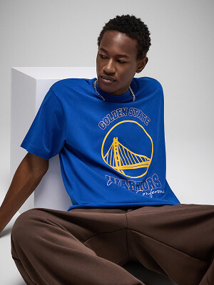 Model v modré košili Golden State Warriors se žlutým logem týmu a hnědých kalhotách na šedém pozadí.