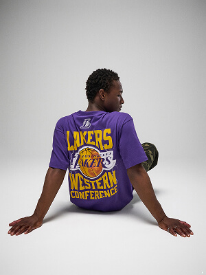 Model ve fialové košili Los Angeles Lakers s velkým žlutým potiskem na zádech a logem týmu, sedí na šedém pozadí zády k fotoaparátu.