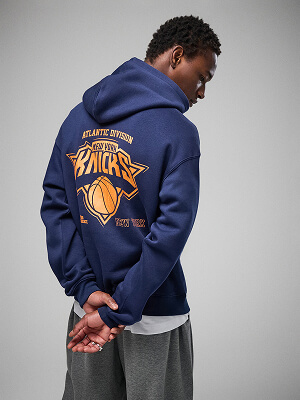 Model v tmavě modré mikině s kapucí New York Knicks s velkým oranžovým logem týmu na zádech a šedých kalhotách na šedém pozadí.